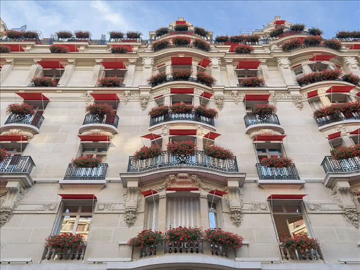 Фасад отеля Plaza Athénée в Париже с красными маркизами и красными цветами на балконах снят с нижнего ракурса